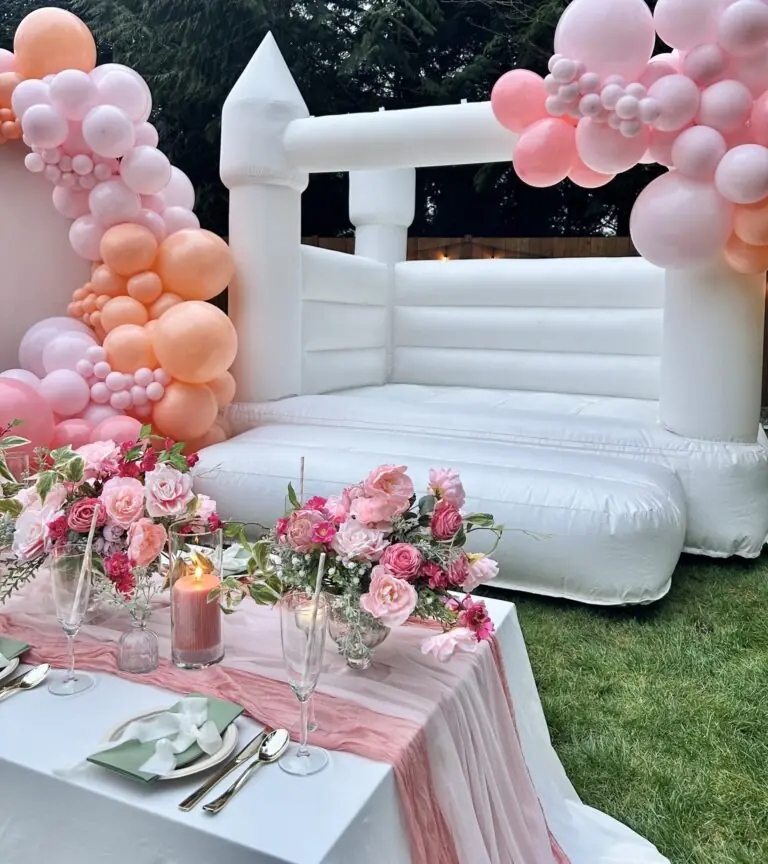 wedding setup with luxury bounce house rentals. Showcasing a white bounce house castle, balloon garlands, backdrop panel, stunning petite floral arrangement tabletop decor for kids seating.