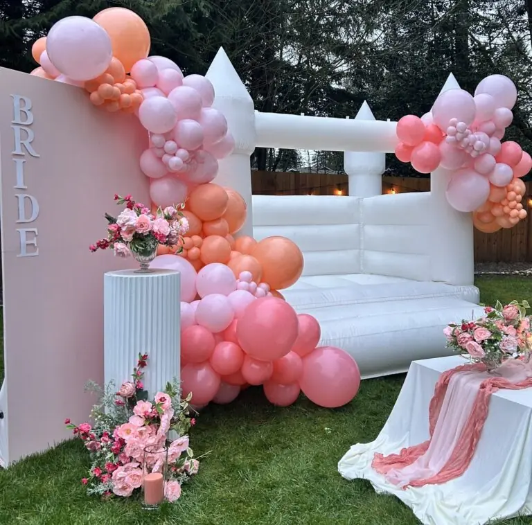 stylish wedding with luxury bounce house rentals, white bounce house castle, balloon garlands, kids seating, floral arrangements tabletop decor, pink backdrop panel with vinyl signage that says "bride" and a custom cake stand with a floral arrangement on it in snohomish county & king county. Bounce House Rental in Marysville