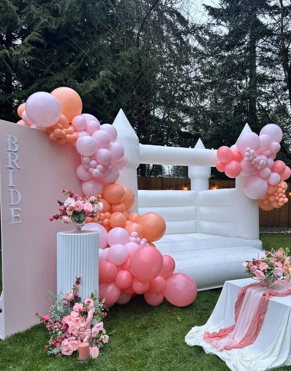 stylish wedding with luxury bounce house rental in arlington, white bounce house castle, balloon garlands, kids seating, floral arrangements tabletop decor, pink backdrop panel with vinyl signage that says "bride" and a custom cake stand with a floral arrangement on it in snohomish county & king county