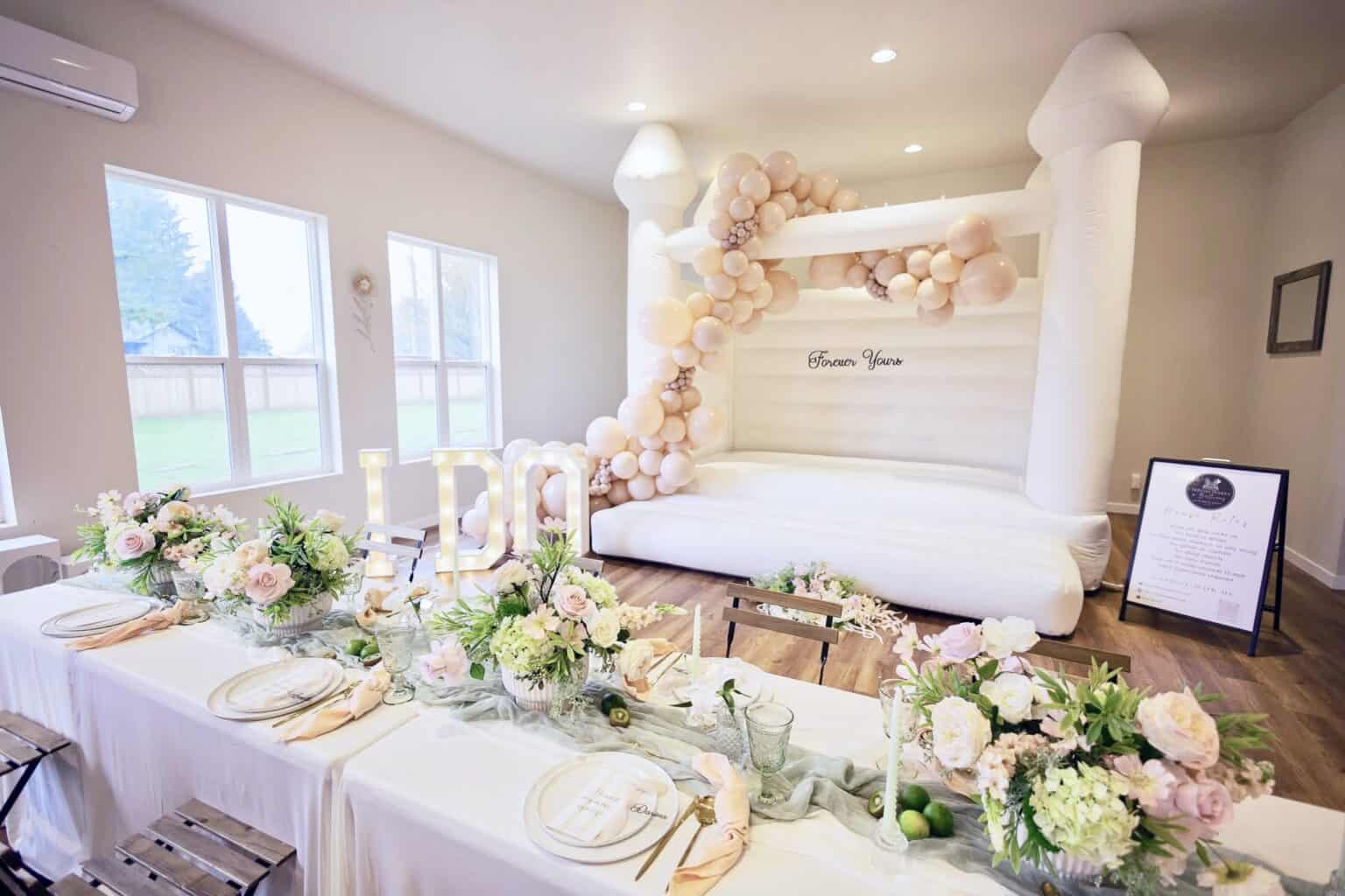 Indoor styled event scene with elegant table florals and a white bounce house in the background; premium party aesthetic.