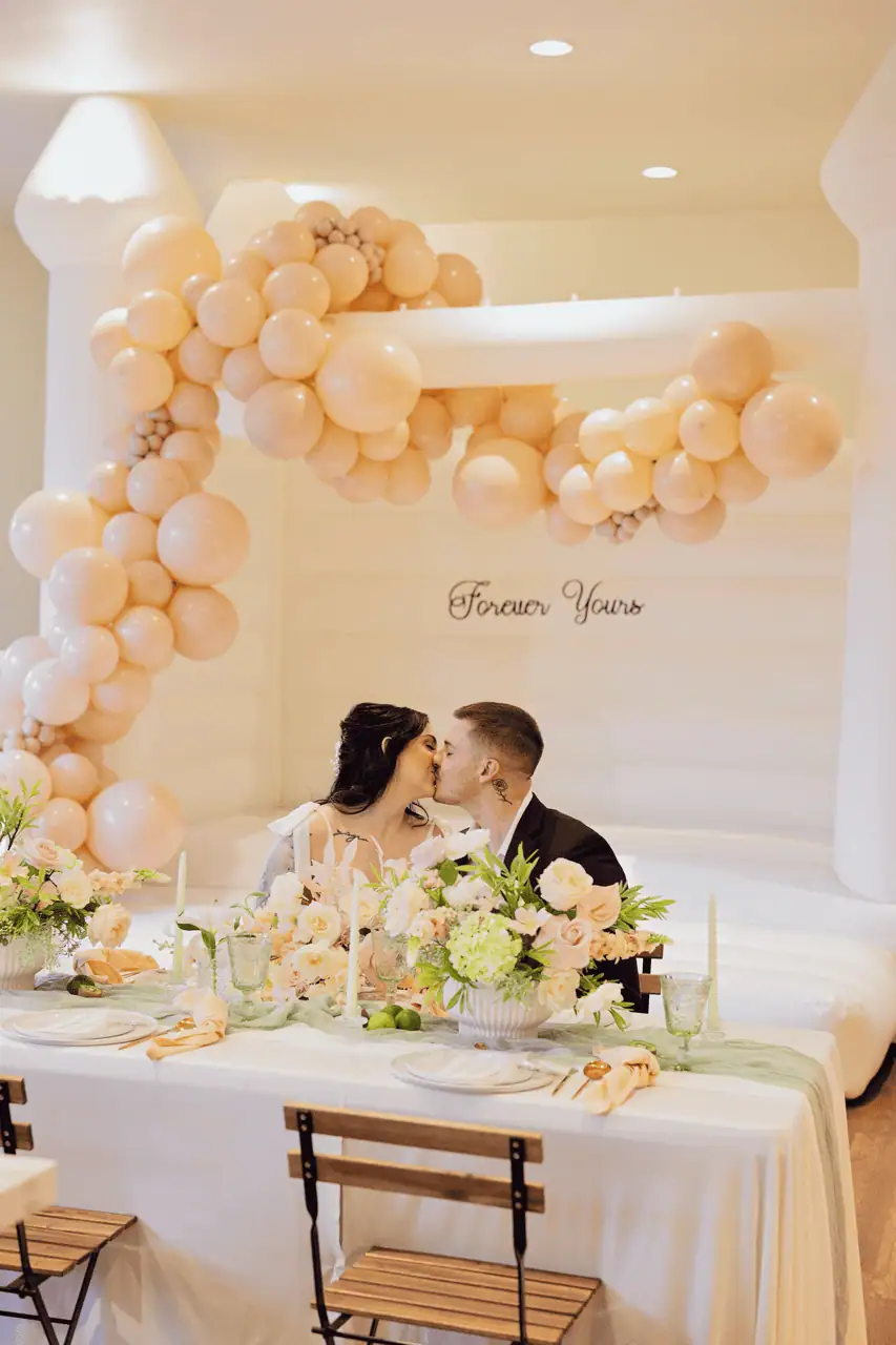 Bride and groom at their table in front of the Magnolia white bounce castle with a cream balloon garland and a Forever Yours sign
