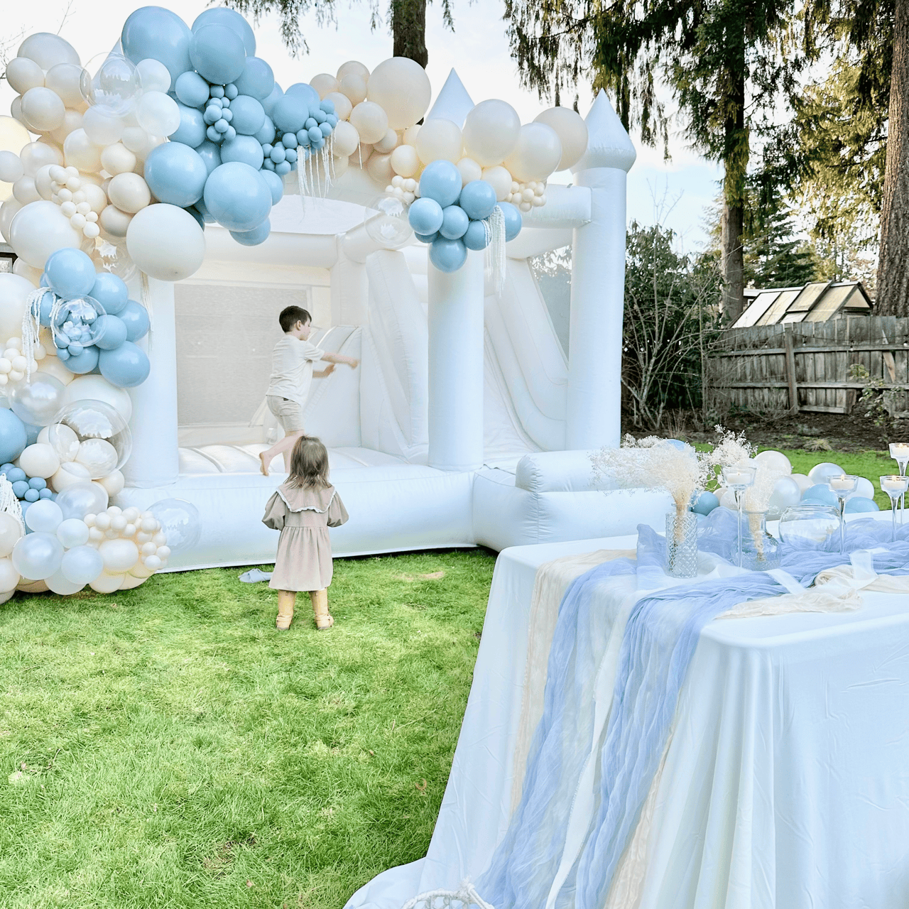 Photo: organic balloon garlands wrapped around a luxury white bounce house at a backyard event