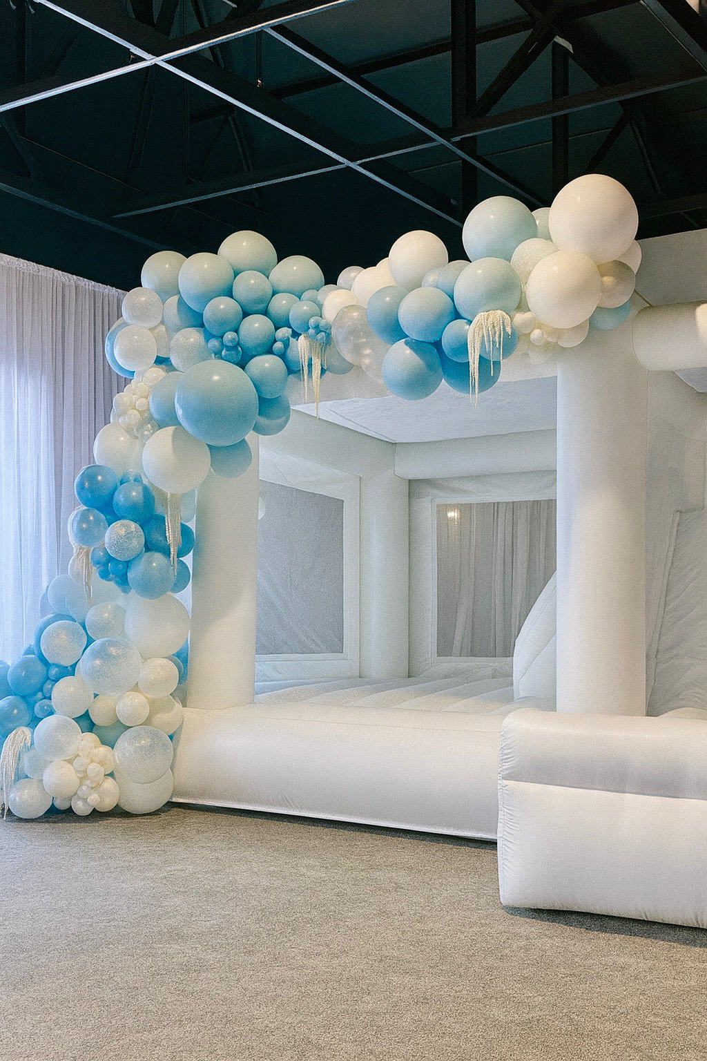 Photo: Winter Wonderland balloon garland framing a white bounce house rental in a modern indoor venue.
