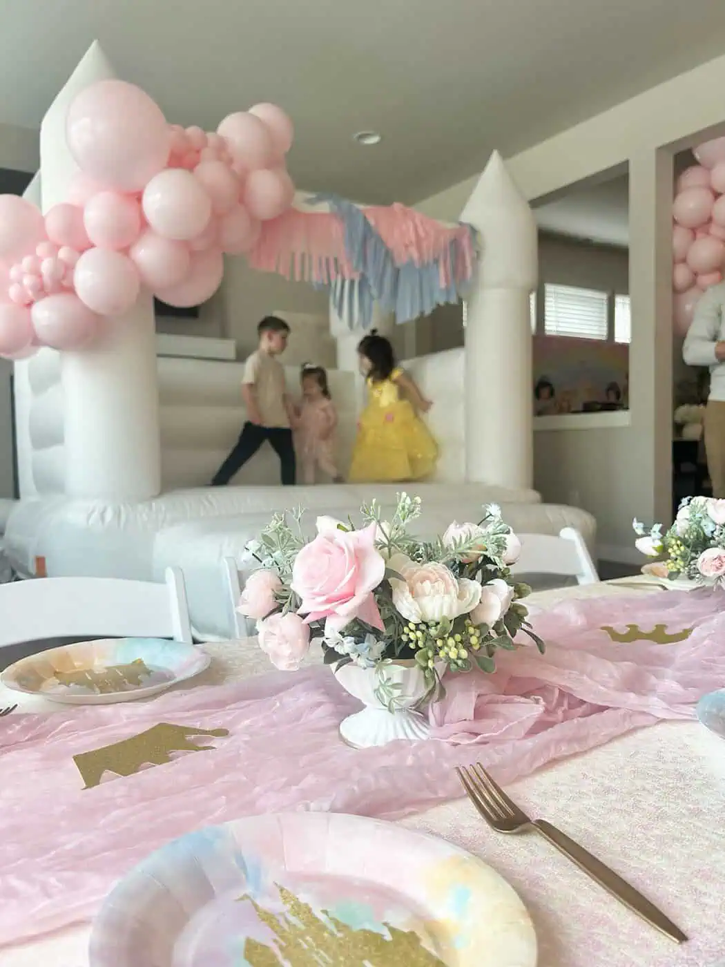 Kids playing in the Thaya white bounce castle indoors behind a princess party table with pink roses and a blush runner