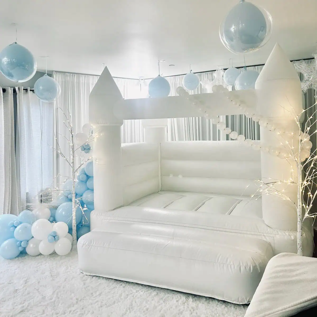 White bounce castle styled for a Winter Wonderland party with pale blue and white balloons, hanging orbs, a pearl strand and lit branches